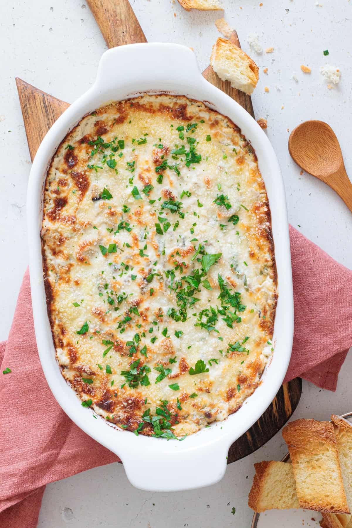 Close-up overhead shot of golden baked creamy mushroom dip in a white oval dish garnished with fresh parsley, surrounded by toasted crostini