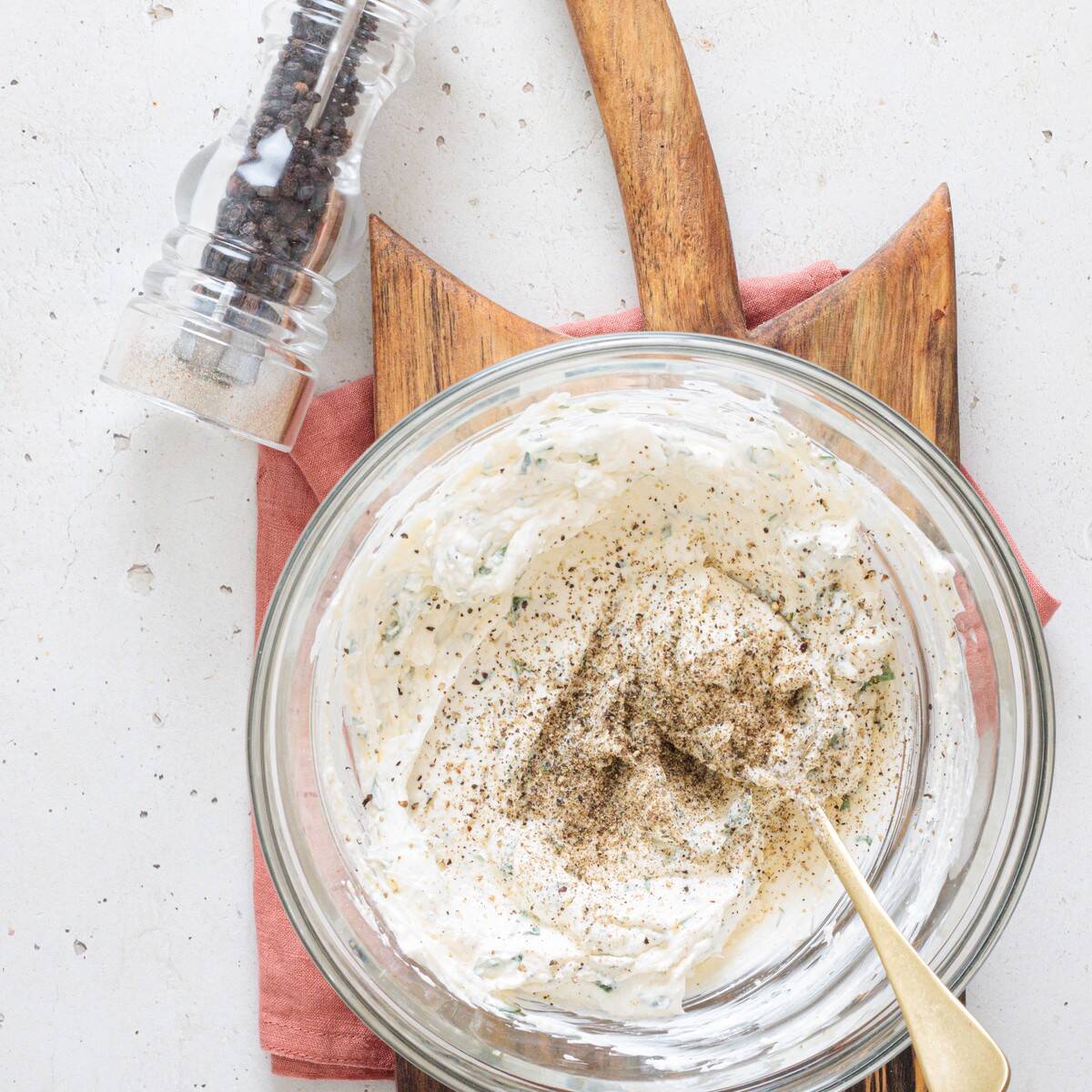Creamy mushroom dip base in a glass bowl being seasoned with freshly ground black pepper, with a pepper grinder in the background