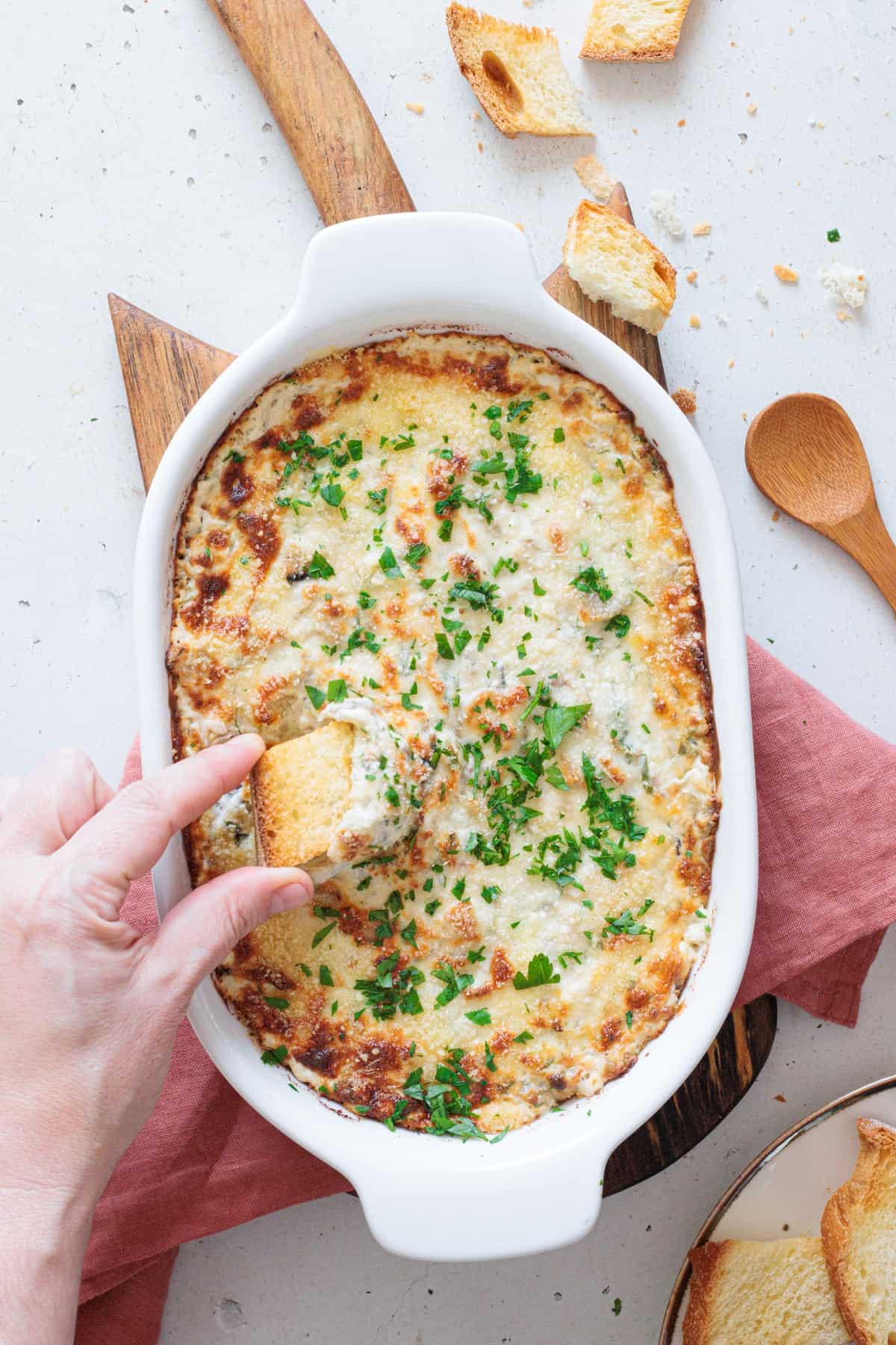 A hand scooping a piece of toasted crostini into golden baked creamy mushroom dip straight from the white oval baking dish