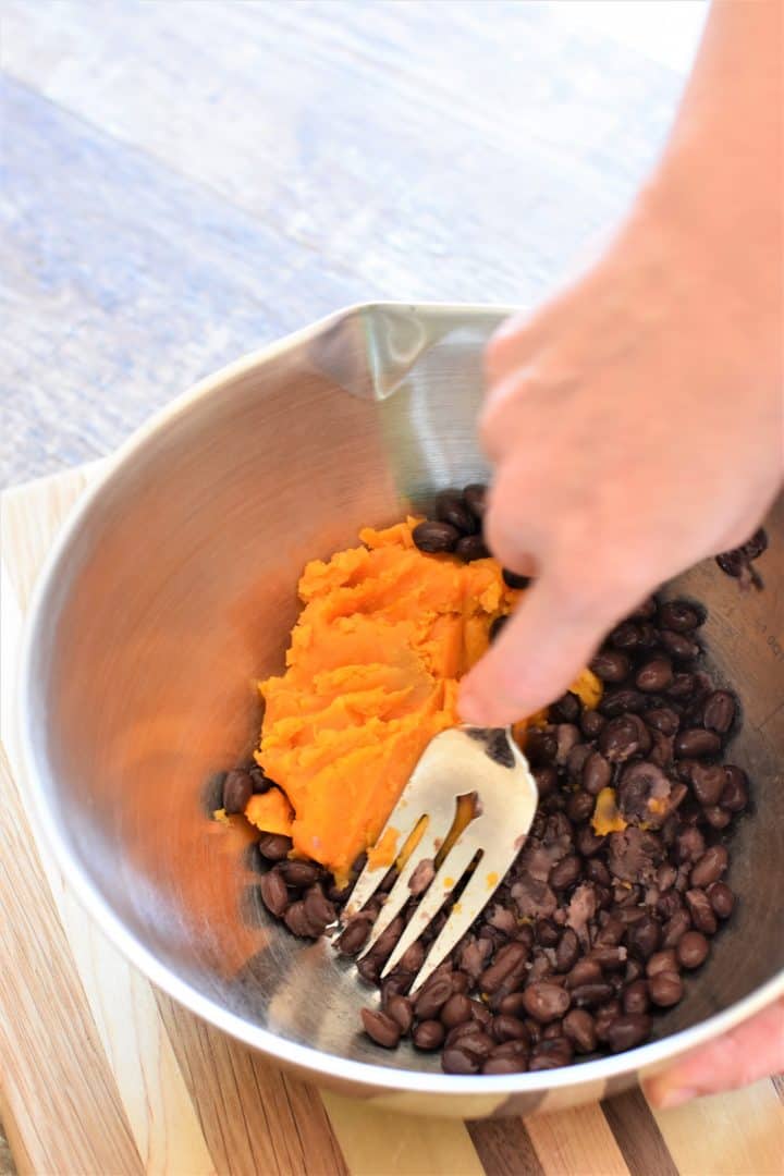 Mashing black beans and sweet potato together with a fork in a mixing bowl