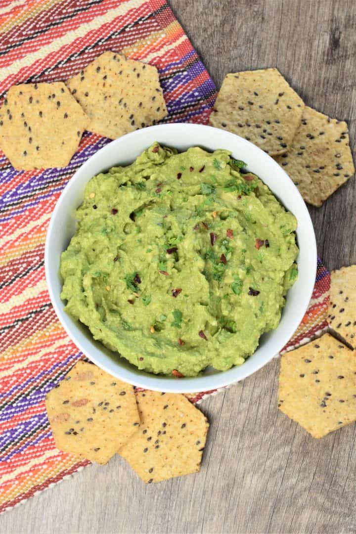 overhead of guacamole in a white bowl on a colorful placemat with tortilla chips around it 