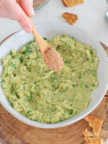 Adding homemade guacamole seasoning from a spoon over a bowl of fresh homemade guacamole