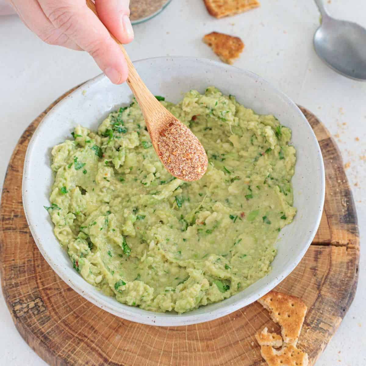 Adding homemade guacamole seasoning from a spoon over a bowl of fresh homemade guacamole