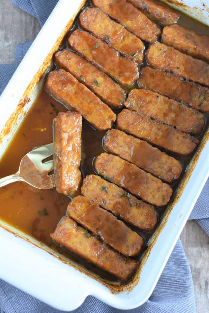 Tempeh in baking dish with serving fork ready to serve one and with some missing
