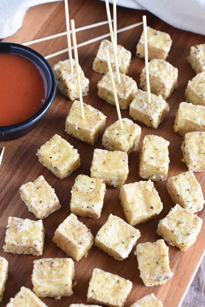 Tofu on wooden serving block with some serving toothpicks in a few pieces and dipping sauce on the side