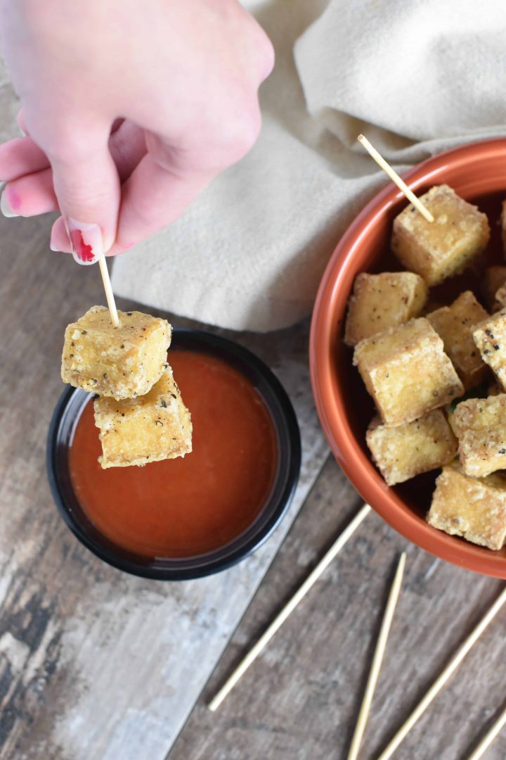 Two pieces of tofu on a serving toothpick about to dip into some sweet chili dipping sauce with the rest of the tofu in a bowl