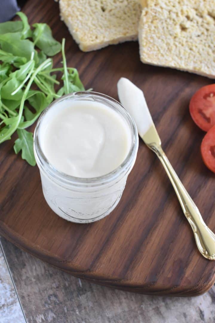 mayonnaise in mason jar with spreading knife next to it and arugula, bread and tomatoes partially in view