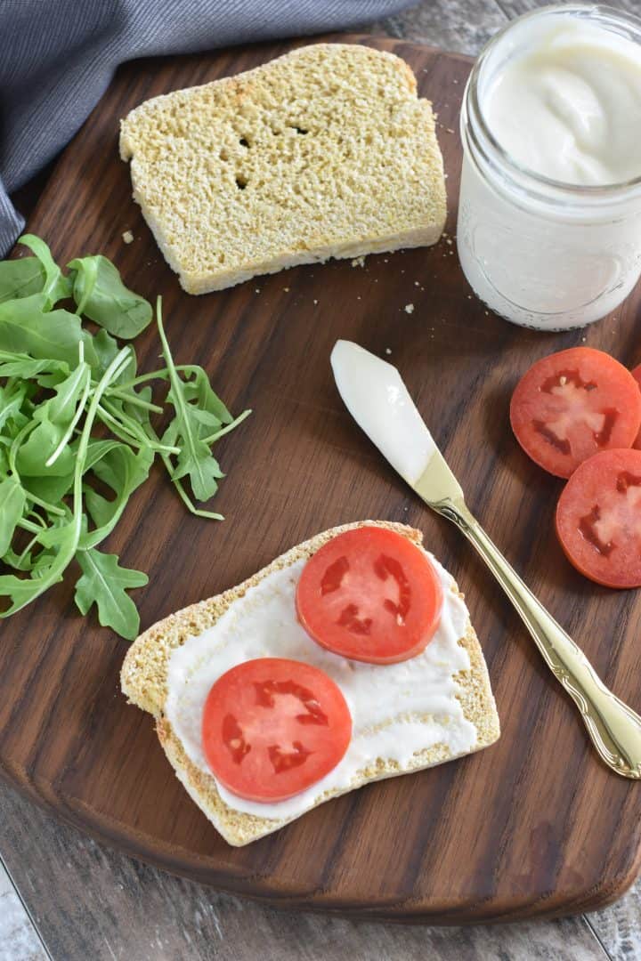 mayo spread on bread with two tomatoes on top, spreading knife next to it and rest of mayo, another piece of bread, arugula and more tomatoes on wooden board