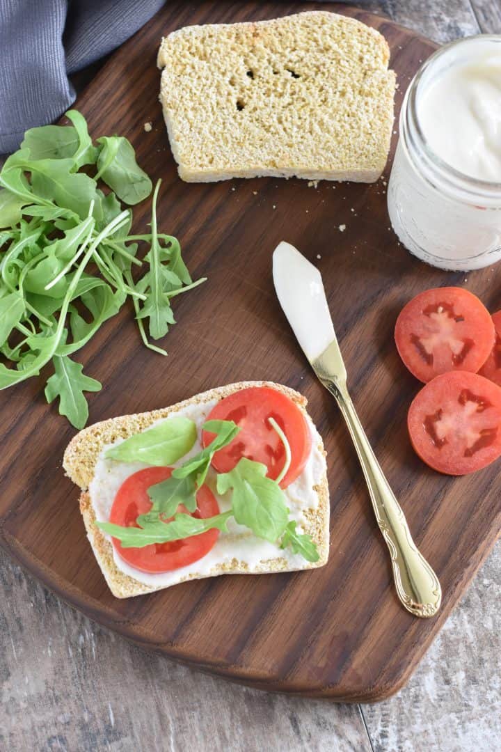 Piece of gluten-free bread with vegan mayo, tomatoes and arugula with spreading knife next to it and more of each item in the background
