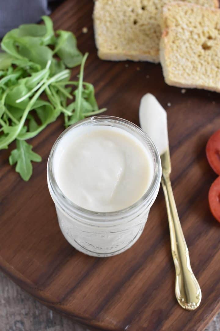 Oil-Free Vegan Mayo in mason jar with knife with some on it next to it and bread, arugula and tomatoes in background