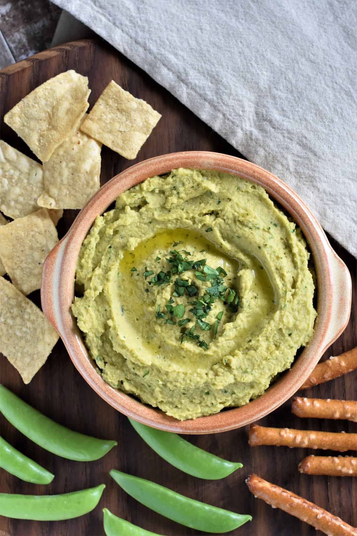 Avocado Hummus in a serving bowl surrounded by tortilla chips, pretzels and sugar snap peas