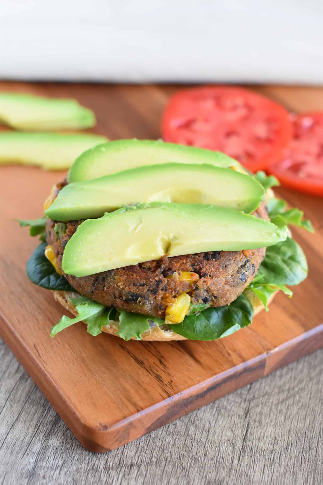 burger on bun with lettuce topped with sliced avocado on a wooden board with avocado and tomato slices in the background