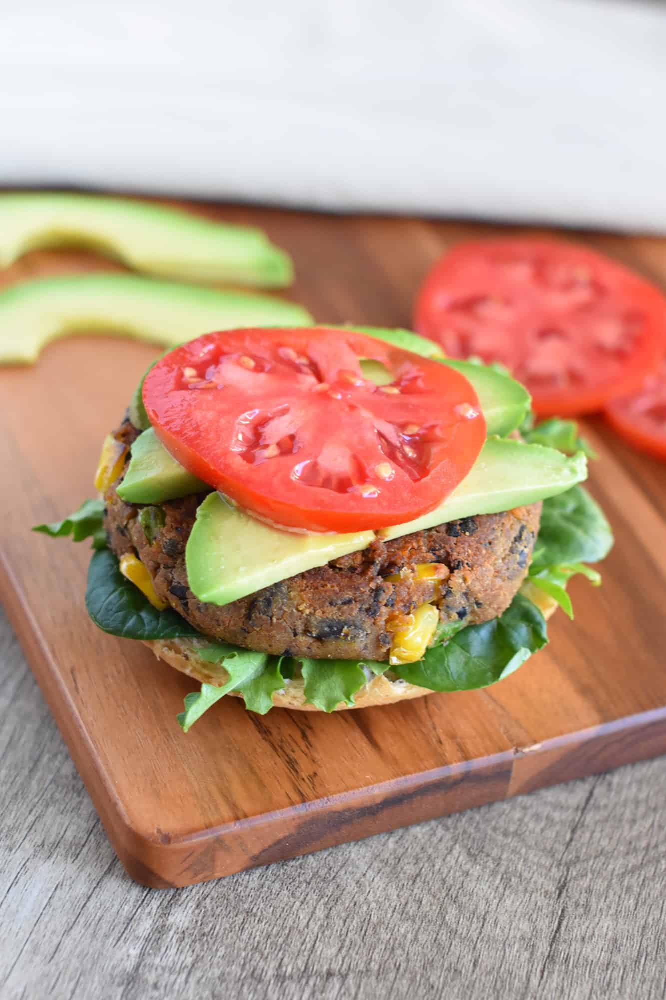 burger on a but with lettuce topped with avocado and tomato on a wooden board with more of the toppings in the background