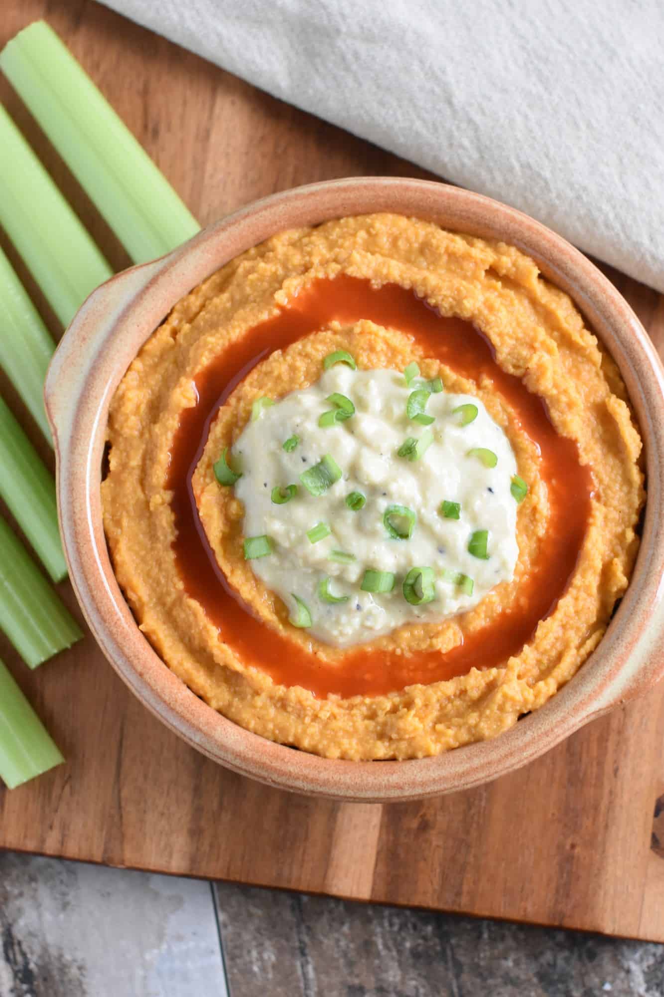 Buffalo hummus in a serving dish on a wooden board with celery sticks next to it
