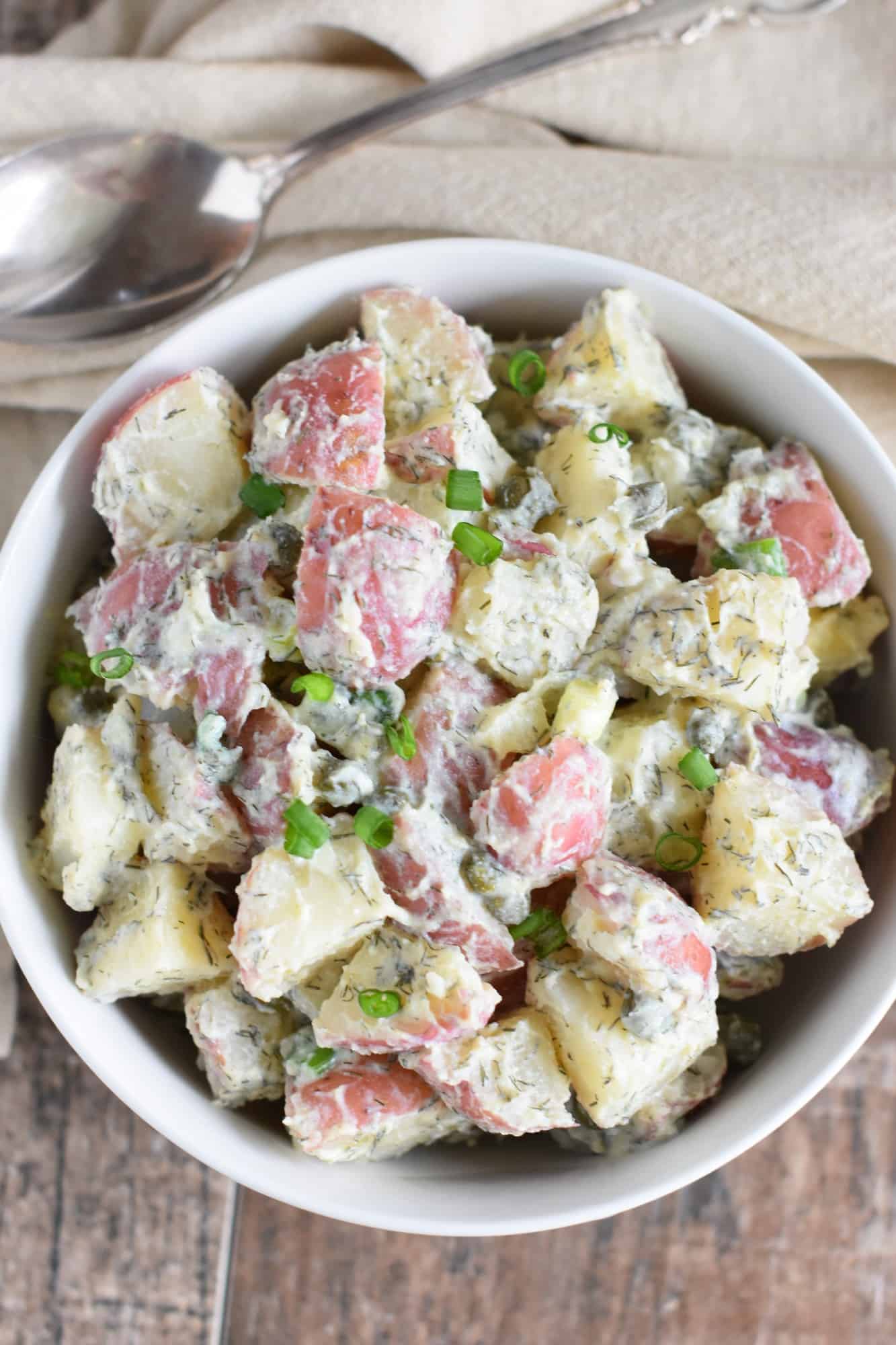 overhead of potato salad in a white bowl with tan napkin with spoon on it behind it
