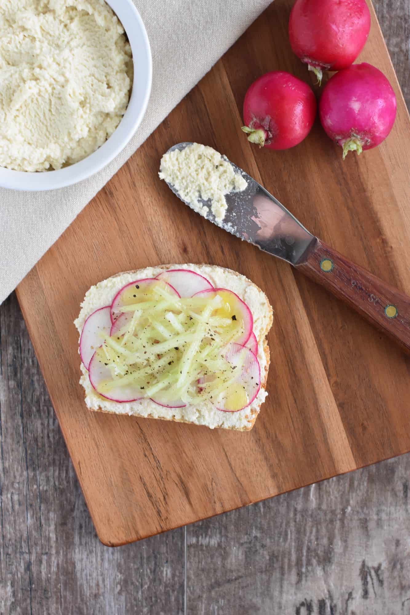 one slice of cucumber and radish ricotta toast with radishes, knife and more ricotta on wooden board next to it