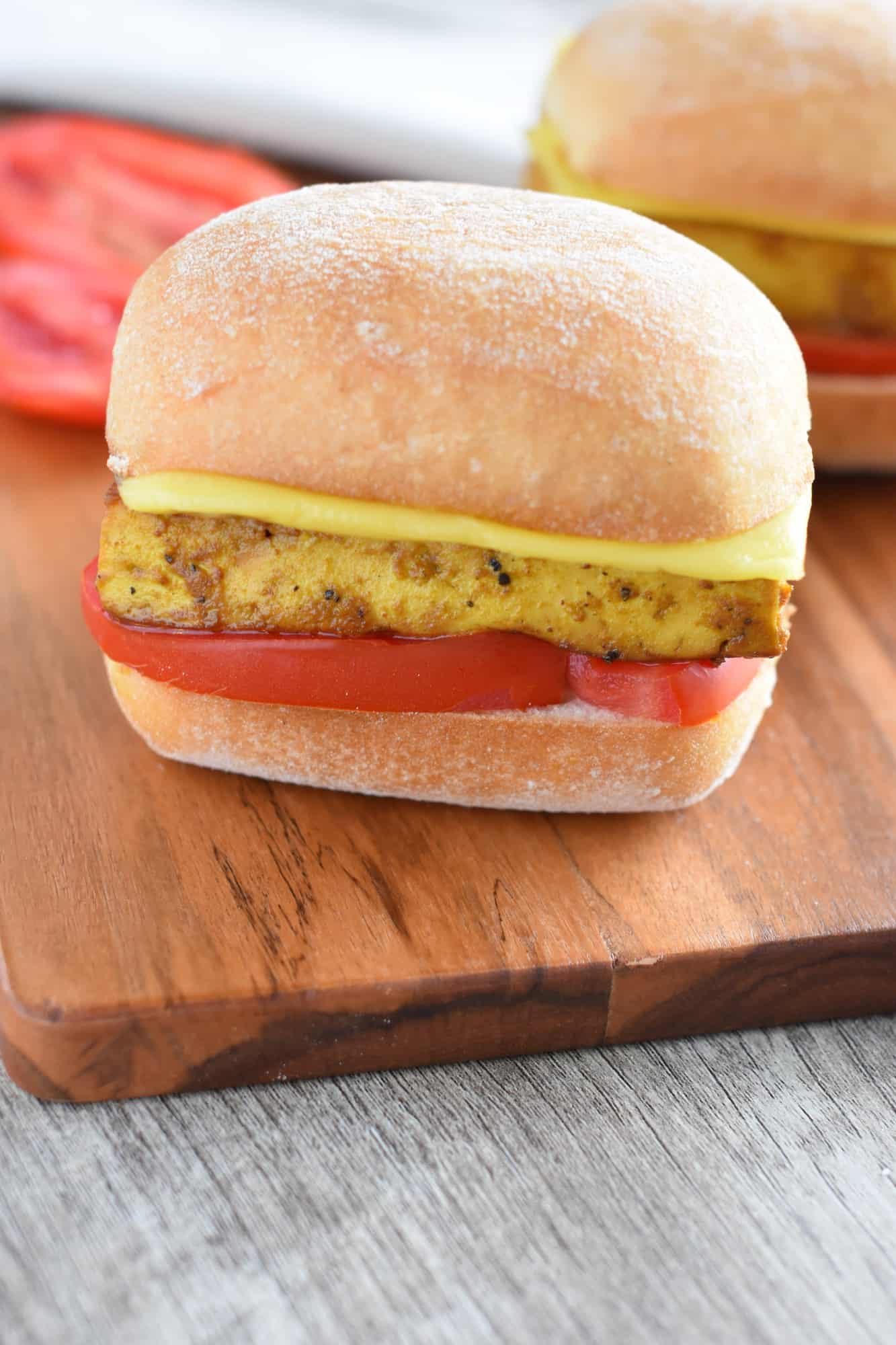 front view of one of a vegan breakfast sandwich on a wooden board with one in the background an a tomato on the board