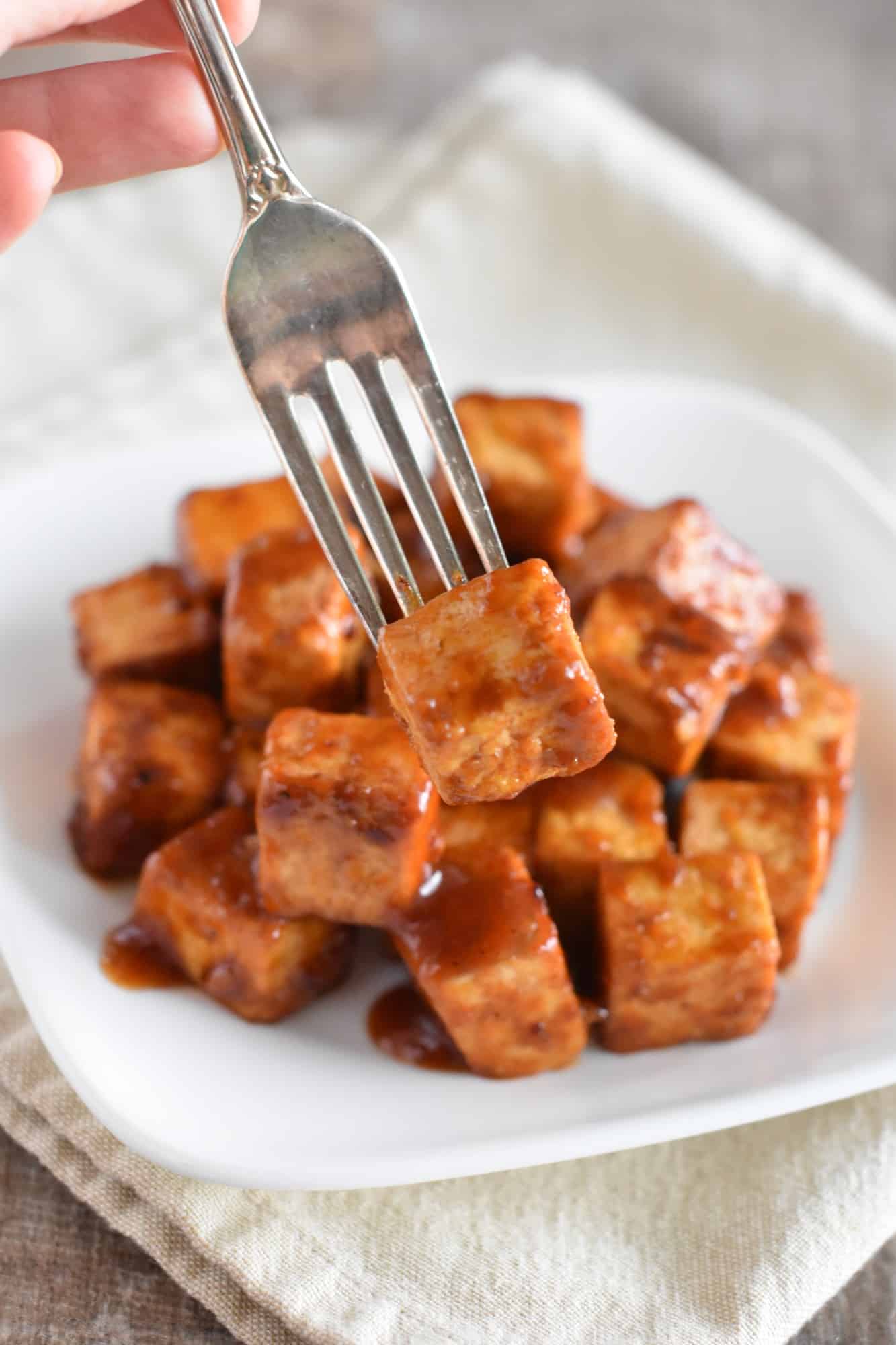 holding a fork with a piece of tofu on it up over a white dish with more tofu on it