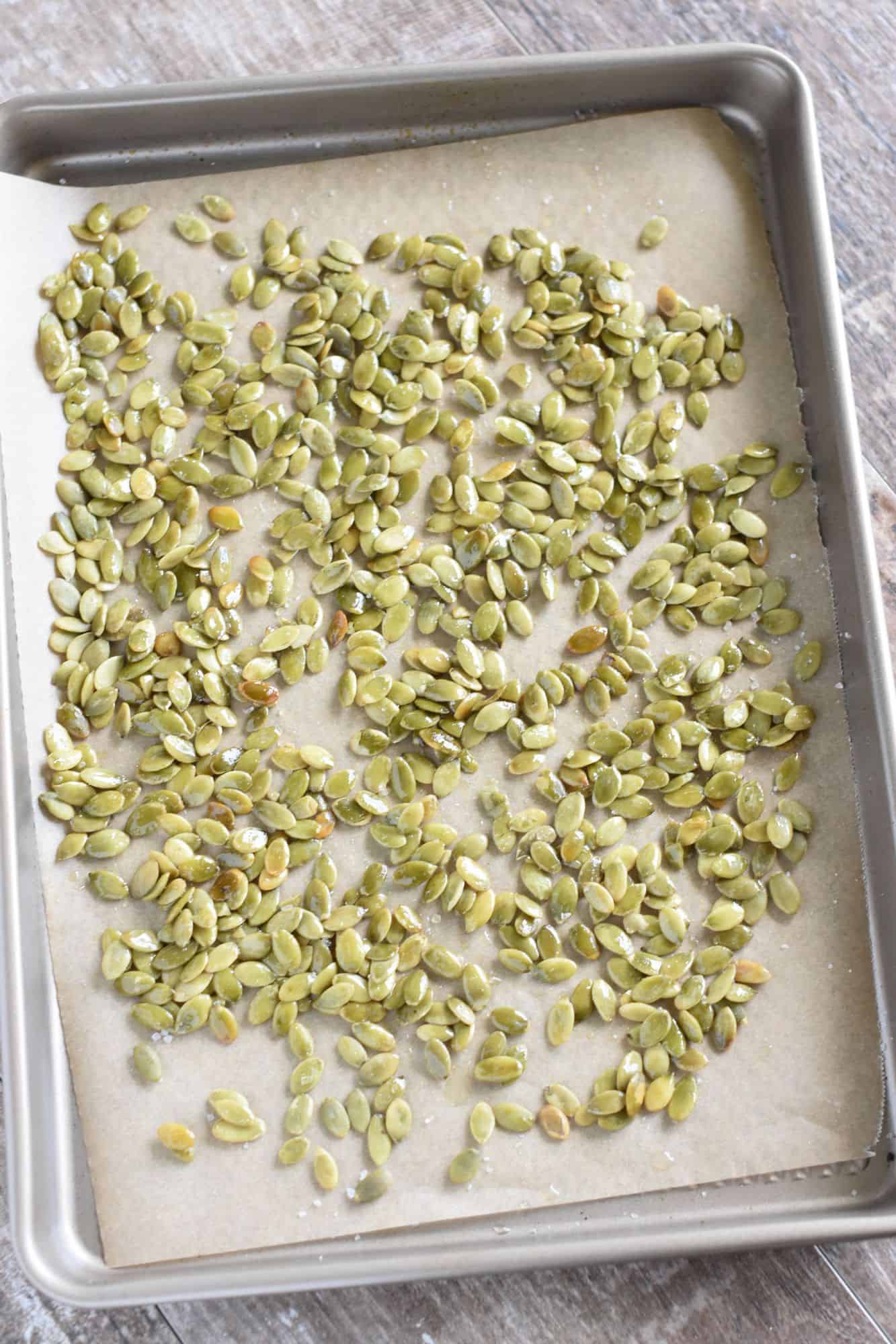 Raw pepitas spread around on the baking sheet after being spritzed with avocado oil and salted