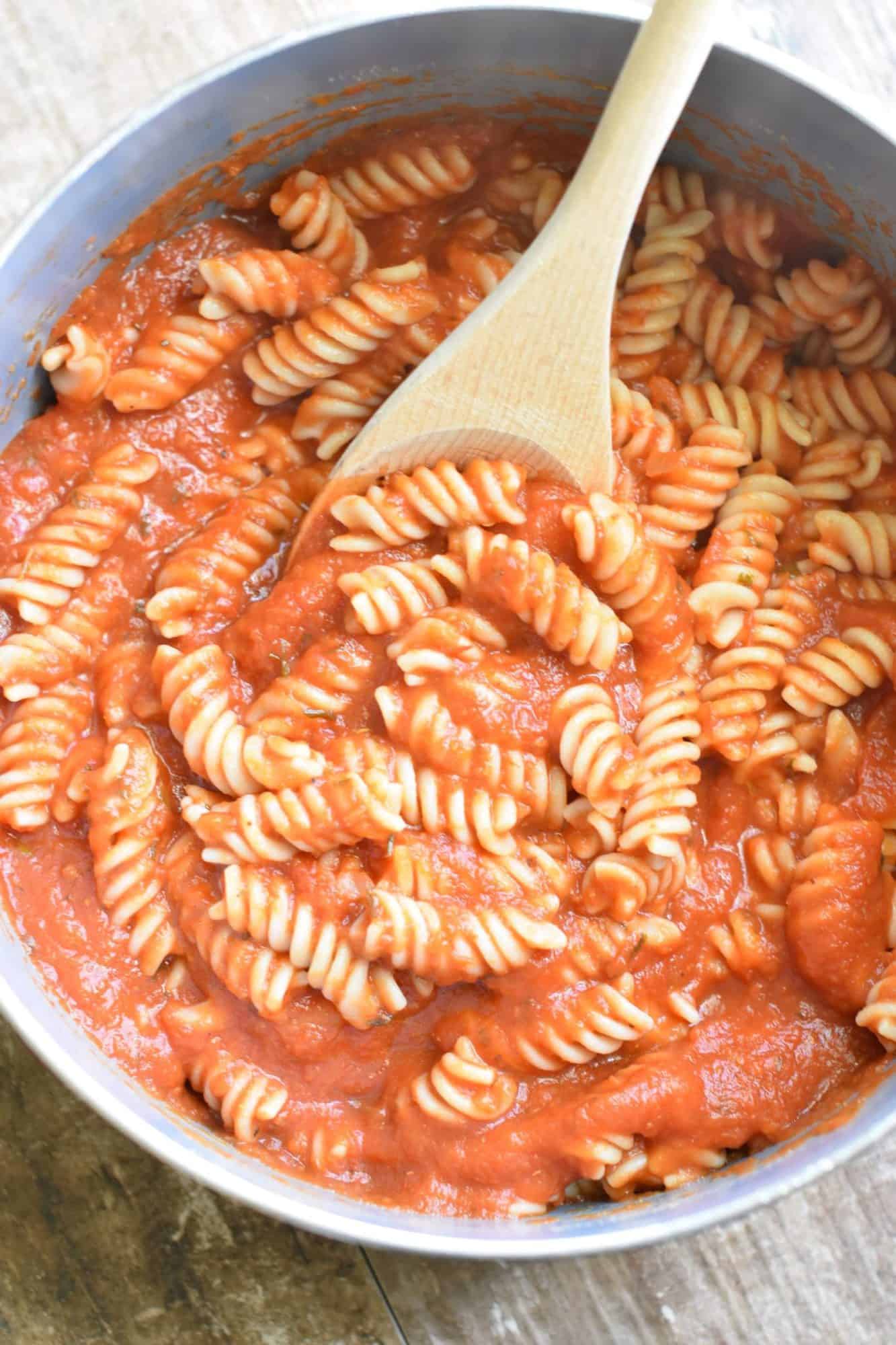 mixing pumpkin tomato sauce and rotini pasta together in a pot with a wooden spoon.