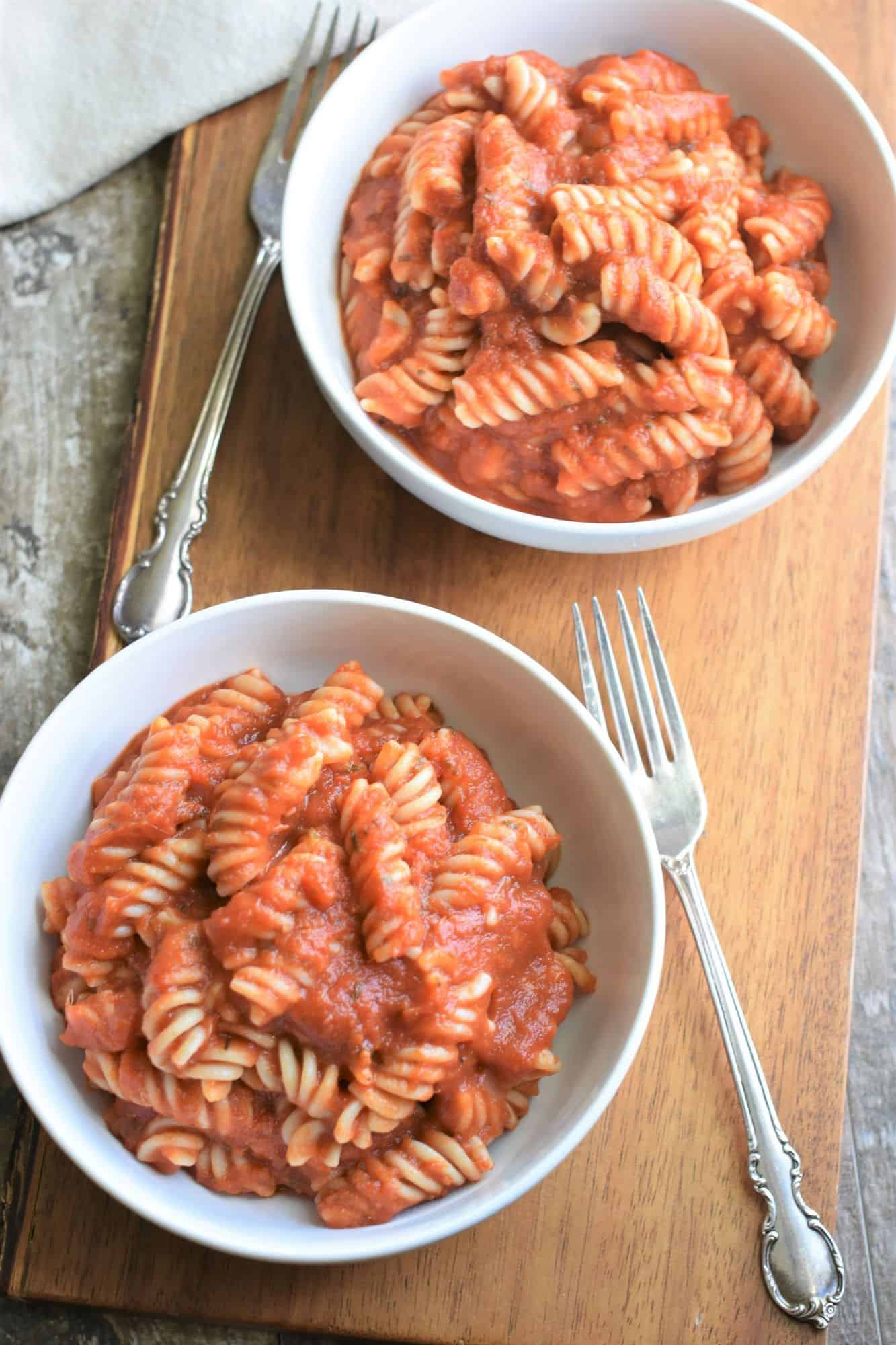 two bowls of pasta with sauce and two forks on a wooden board