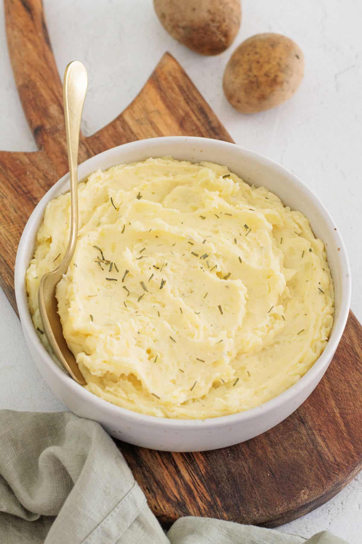 bowl of dairy free mashed potatoes with a golden spoon lying on a wooden board with Yukon potatoes in the background.