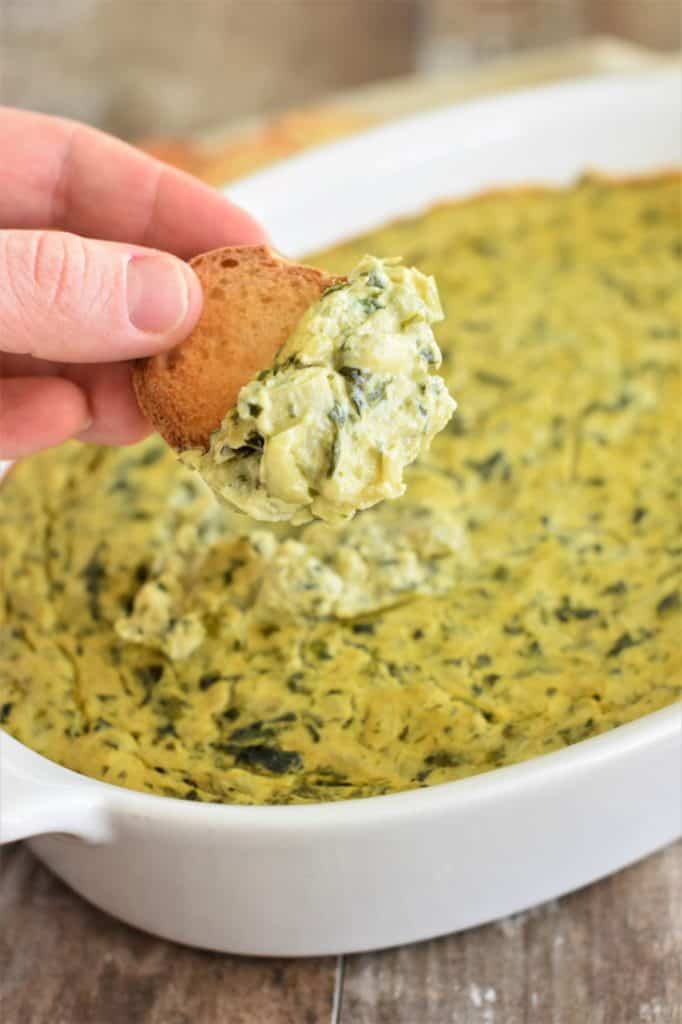 holding a piece of bread with vegan spinach artichoke dip on it over the casserole of the rest of the dip