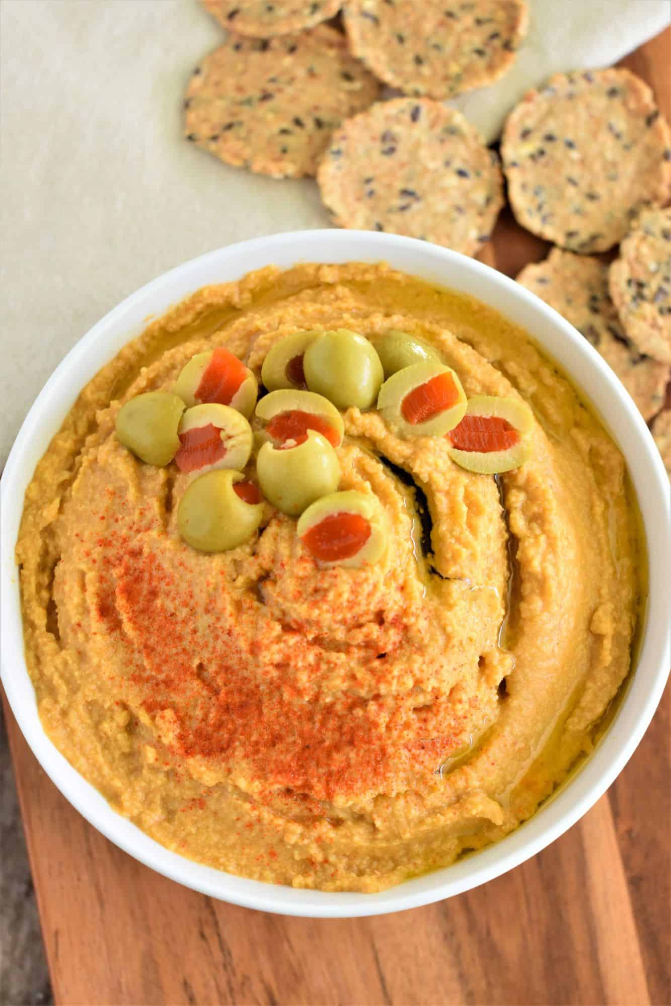 overhead of hummus in white bowl on wooden board with crackers behind it