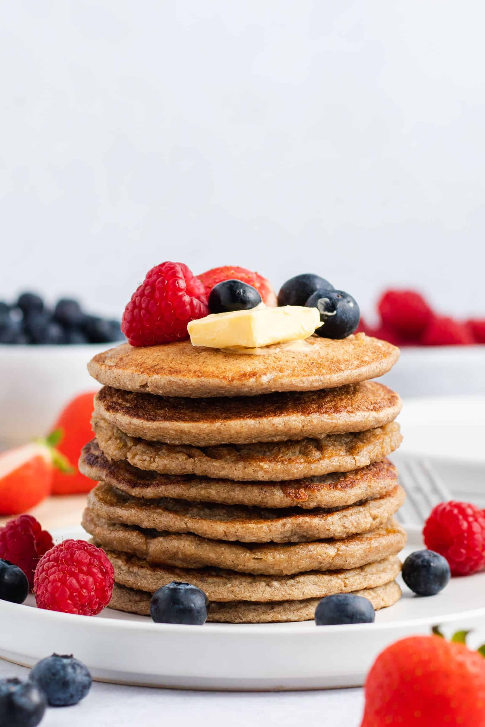 stack of 8 pancakes on a white serving dish with vegan butter, maple syrup and fresh fruit