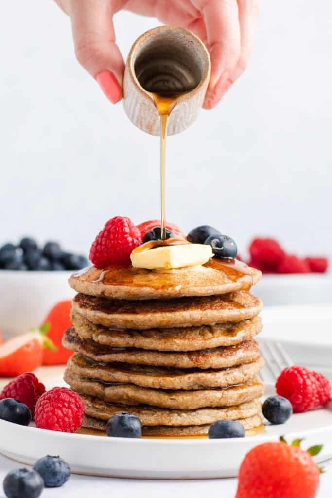 pouring maple syrup onto a stack of pancakes topped with vegan butter and fruit