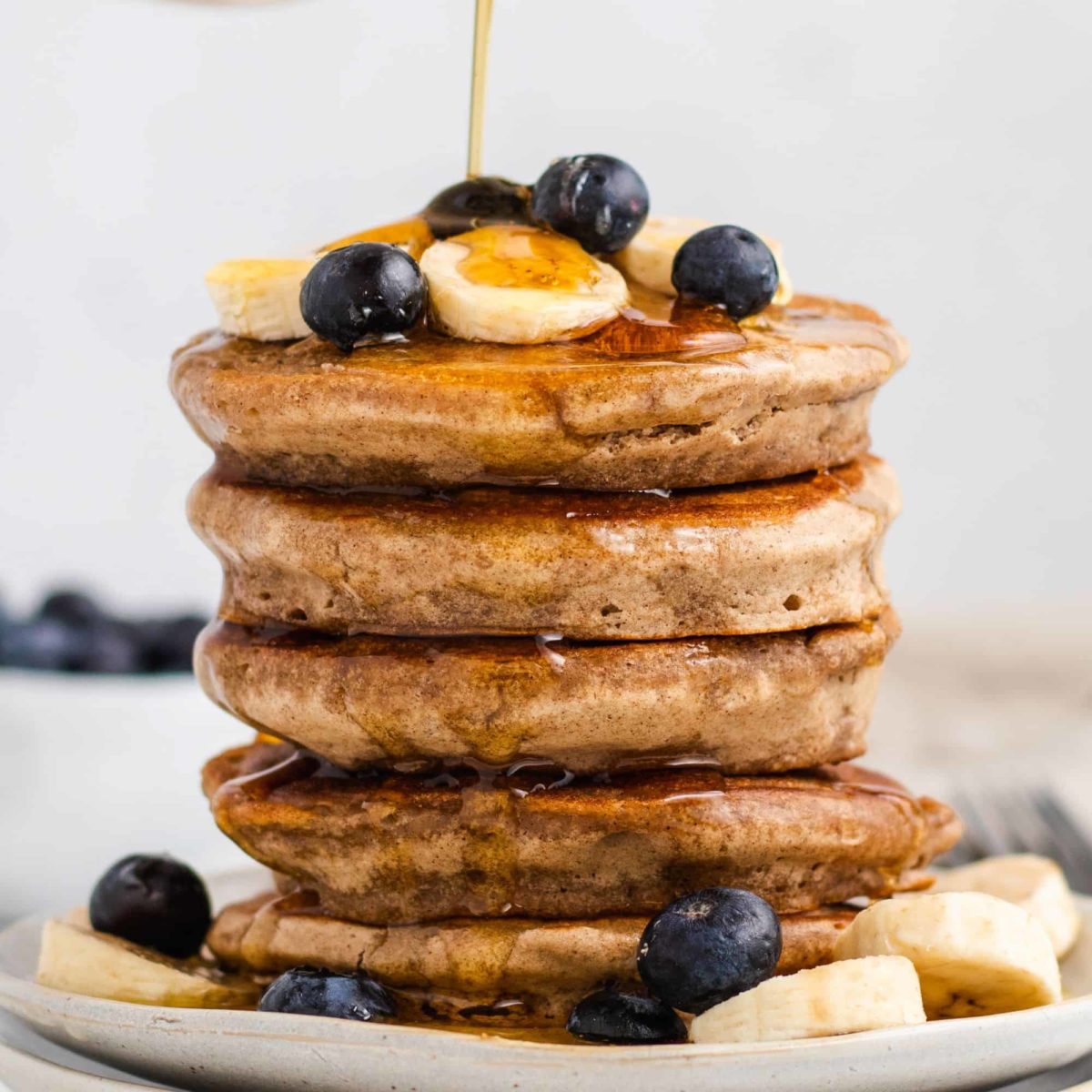 close-up front view of syrup pouring onto stack of pancakes with bananas and blueberries