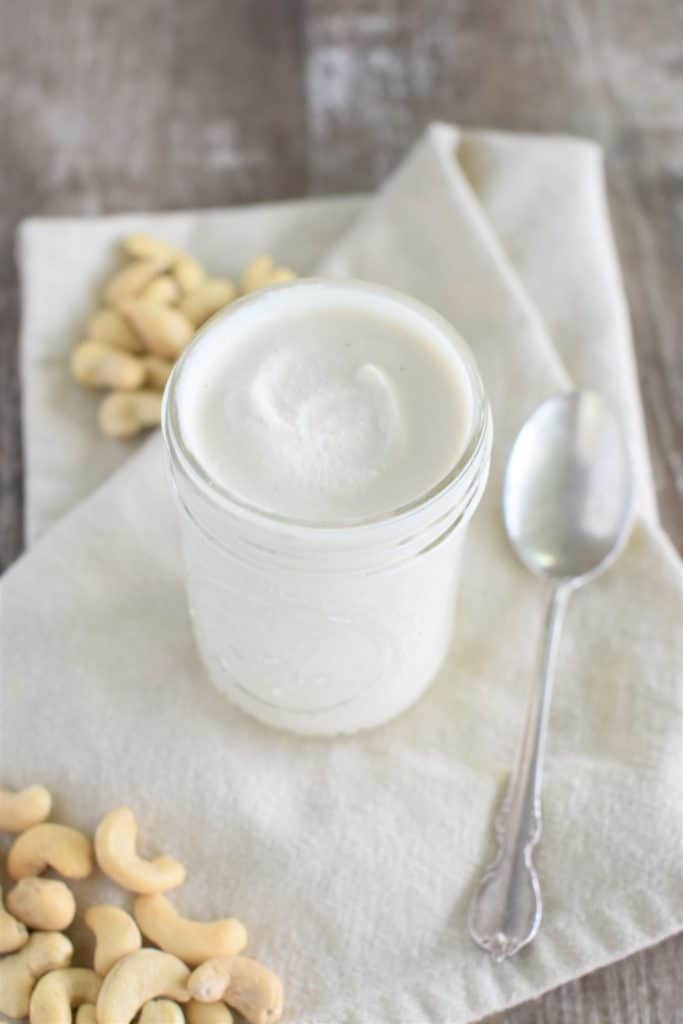 front view of cashew mayo in a mason jar on kitchen towel with some cashews around and a spoon next to it
