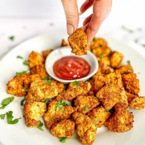 holding a tofu nugget over a plate of more of them with dipping sauce
