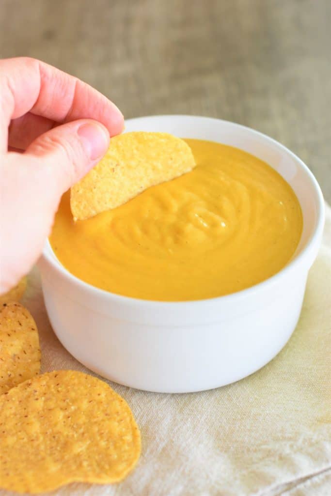 dipping a tortilla chip into the cheese sauce in a white serving bowl