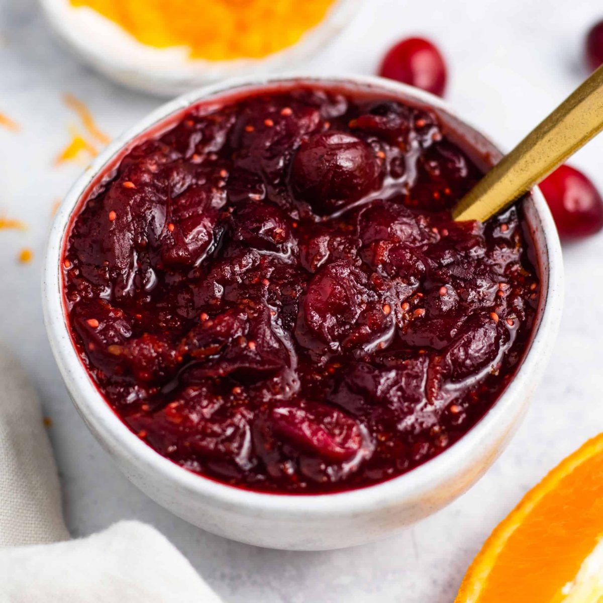 vegan cranberry sauce in serving bowl with spoon in it and some cranberries and orange zest in the background