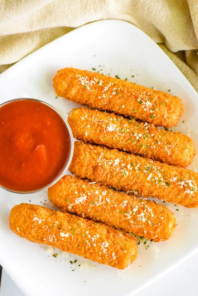 overhead of mozzarella sticks on a white plate with tomato sauce and garnished with Parmesan and parsley