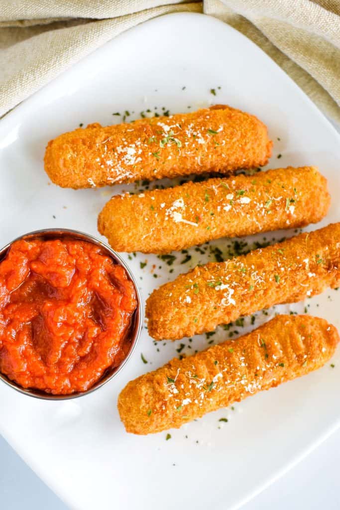 overhead of 4 mozzarella sticks on white plate garnished with Parmesan and parsley with side of marinara sauce