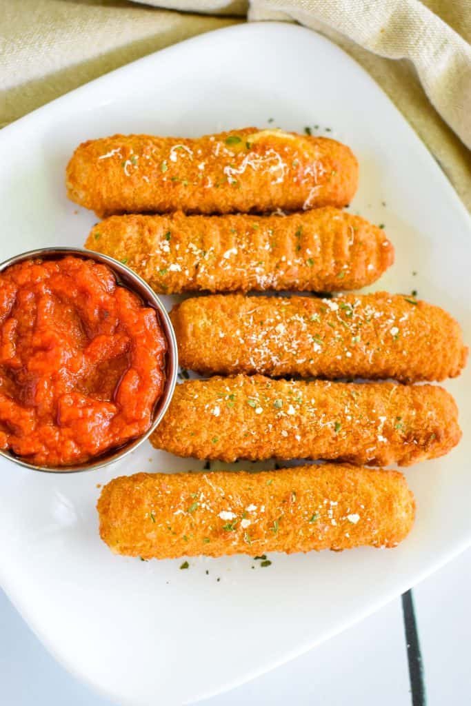 overhead of 5 mozzarella sticks garnished with parsley and Parmesan and cup of sauce on left side of dish