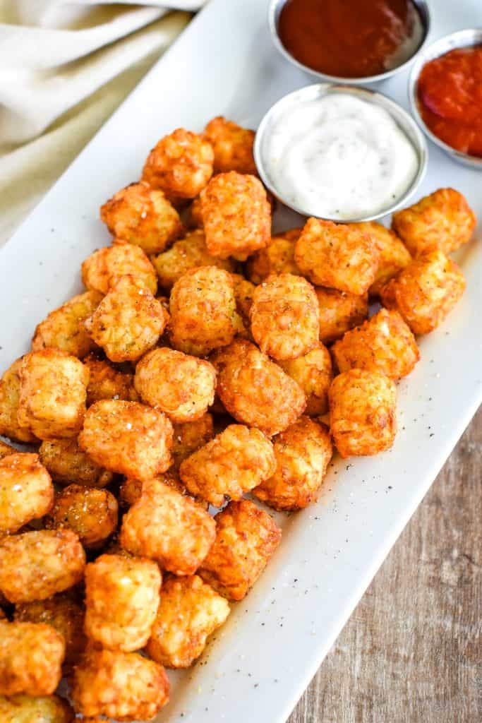 tater tots seasoned with salt and pepper on a white serving plate with three cups of dipping sauces