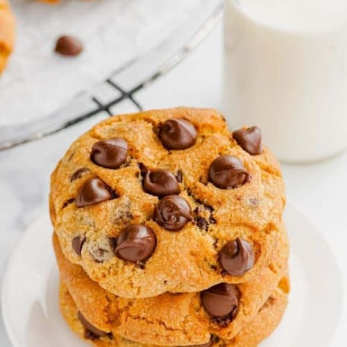 3 stacked chocolate chip cookies on a white plate with some milk and more cookies behind it