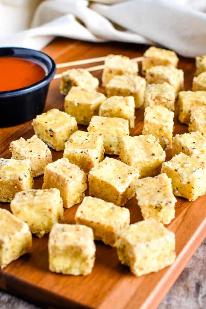 tofu on wooden board with dipping sauce serving toothpicks in the background.