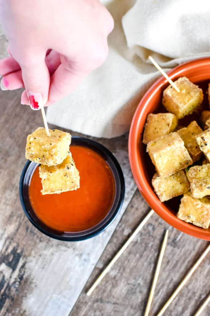 Two pieces of tofu on a serving toothpick about to dip into dipping sauce.