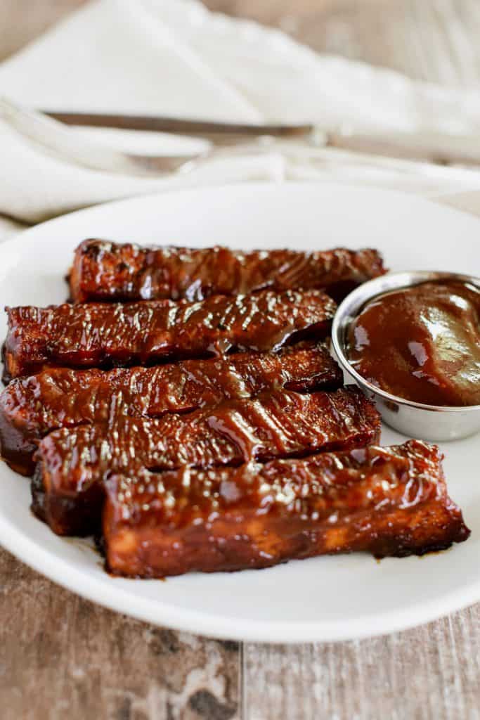 tempeh strips on a white plate with a cup of BBQ sauce next to them.