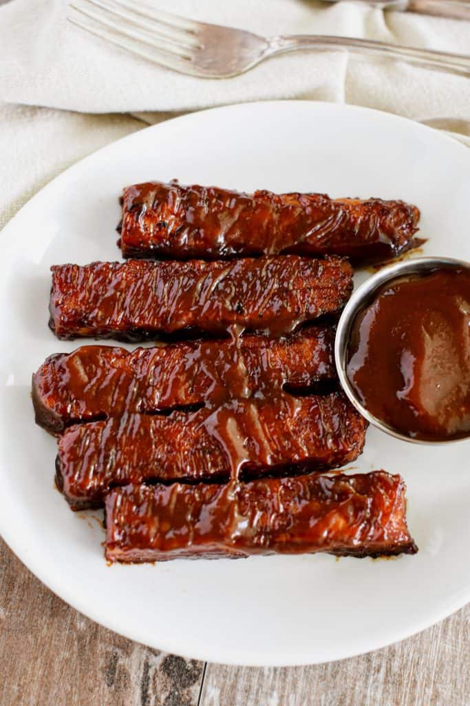 overhead of BBQ tempeh ribs on a white plate with cup of barbecue sauce.