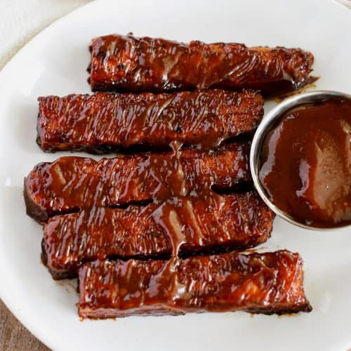overhead of tempeh ribs on a white plate with side of BBQ sauce.