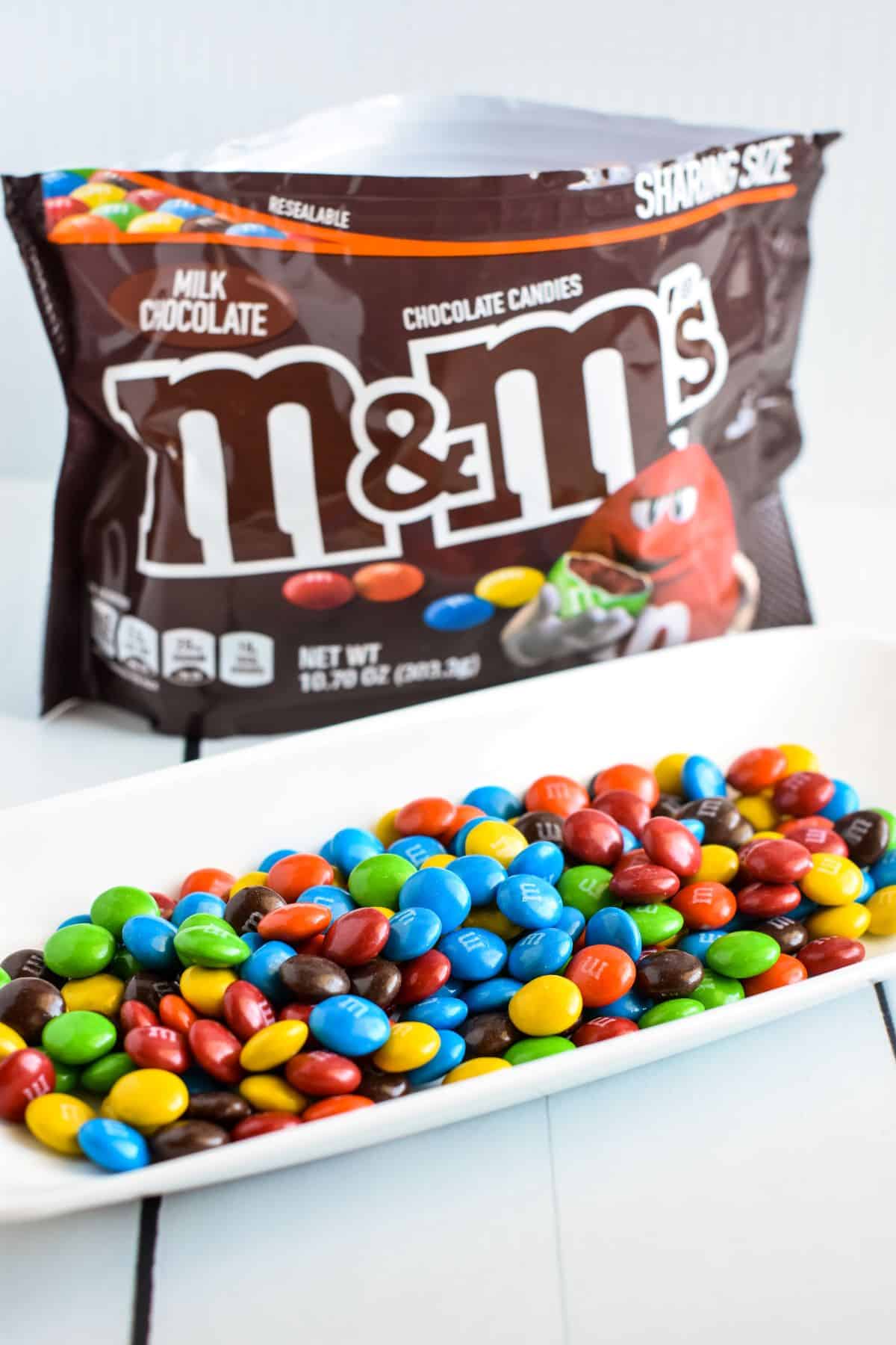 Bag of M&M's in background with M&M's on a white dish in foreground.
