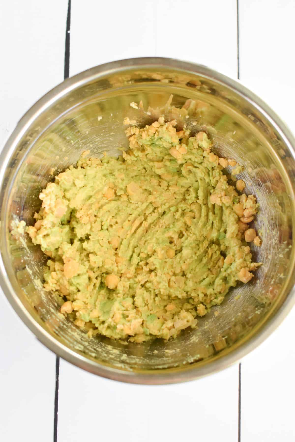 Smashed avocado mixed into chickpeas in a bowl.