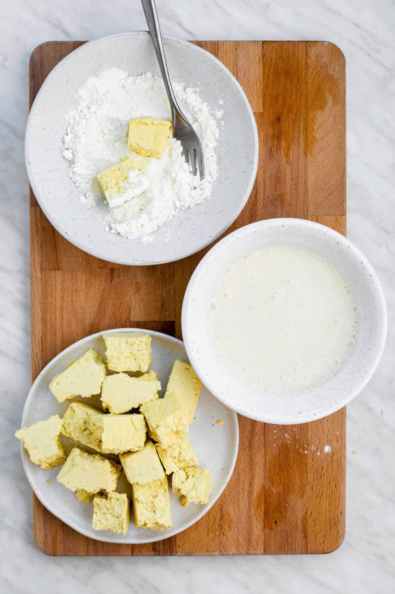 Three white bowls placed on a wooden cutting board. Once filled with corn starch, the second one filled with vegan buttermilk, and the third one with tofu.