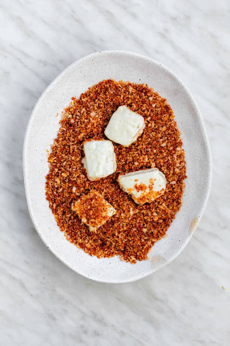 A white plate filled with breadcrumbs and spices. Four tofu pieces placed over the breadcrumbs mixture.