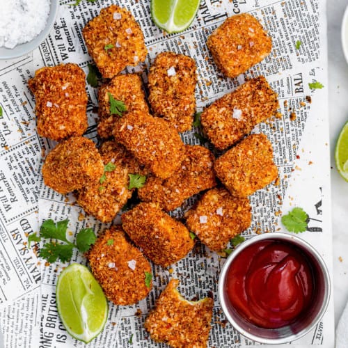 Air fryer tofu nuggets placed on a platter with news paper. Lime edges and a bowl of ketchup placed around them.